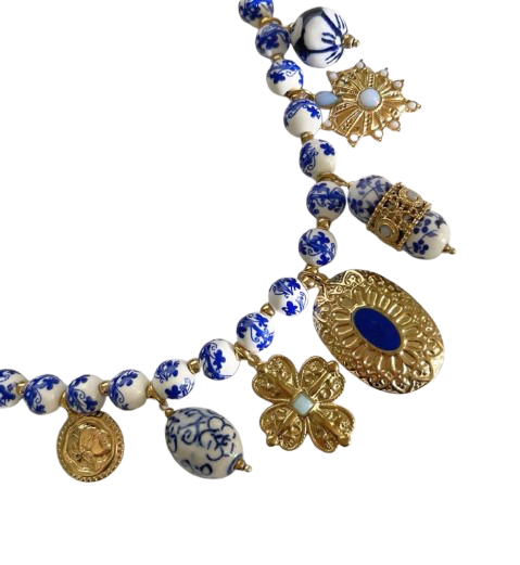 A beaded necklace featuring white ceramic beads with blue floral patterns, gold-toned filigree charms, and a central oval medallion with a deep blue inlay, set against a plain white background.