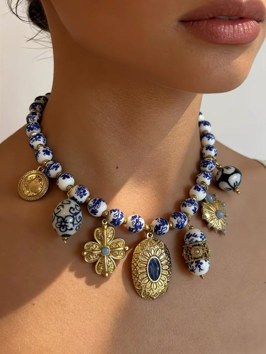 A close-up of a model wearing a statement necklace featuring blue and white patterned ceramic beads, gold filigree charms, and a central oval pendant with a dark blue stone.