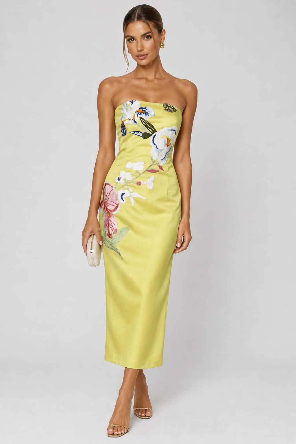 A woman models a strapless yellow midi dress with colorful floral and bird embroidery, standing against a light gray studio background.