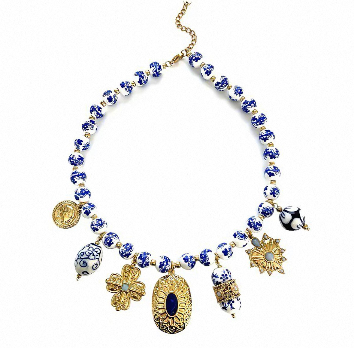 A statement necklace featuring blue and white patterned glass beads on a gold-tone chain, adorned with multiple dangling charms including a gold coin, a blue-and-white floral piece, a gold sunburst, a penguin, and a large oval medallion with a dark blue center, all displayed on a clean white background.